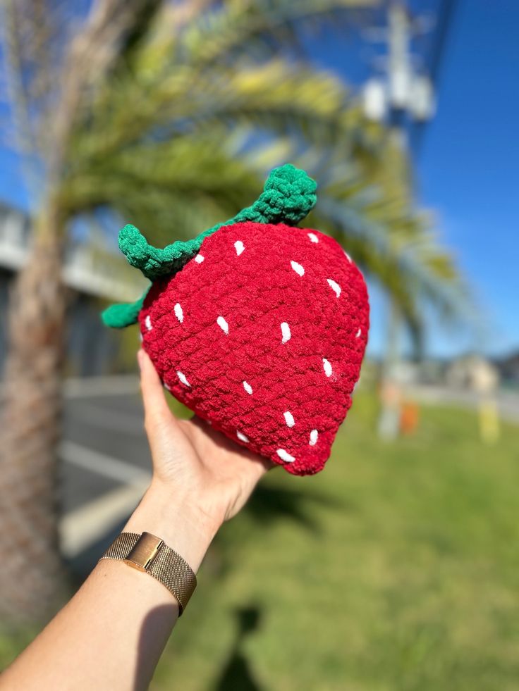 a hand is holding up a knitted strawberry ornament in front of a palm tree