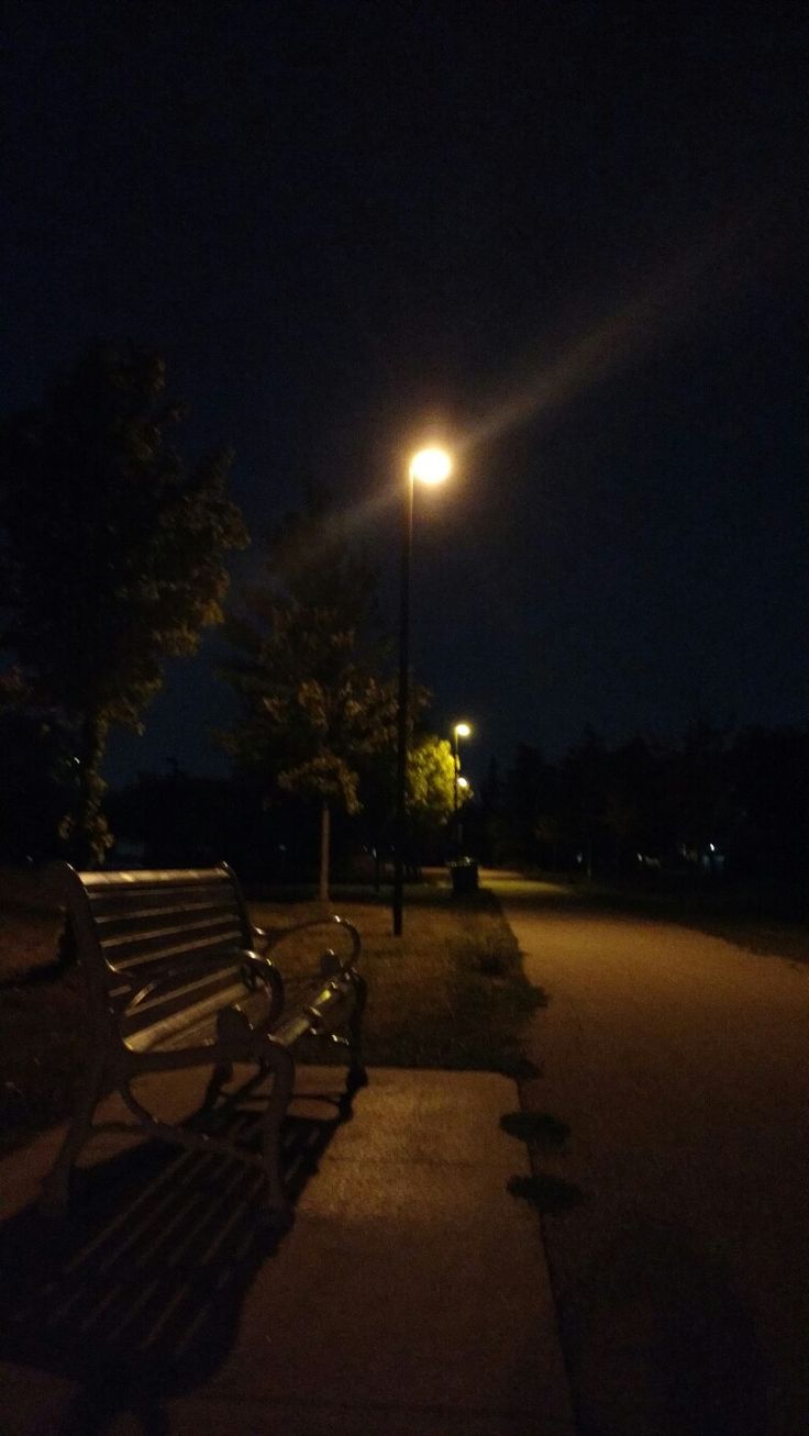 two park benches sitting next to each other on a sidewalk at night with street lights in the background