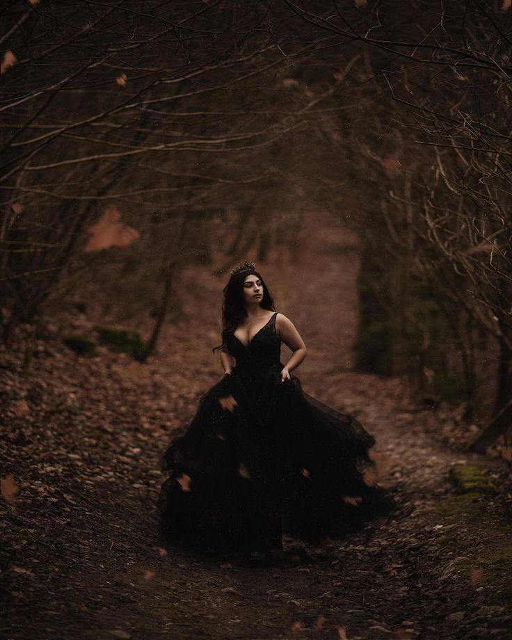 a woman in a black dress is walking through the woods with her hands on her hips