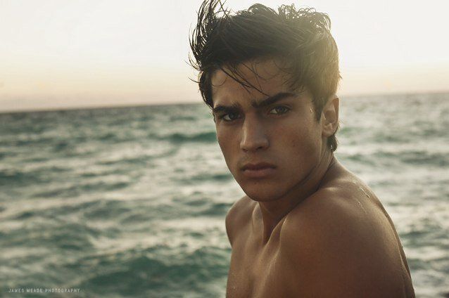 a shirtless young man standing in front of the ocean with his head turned to the side