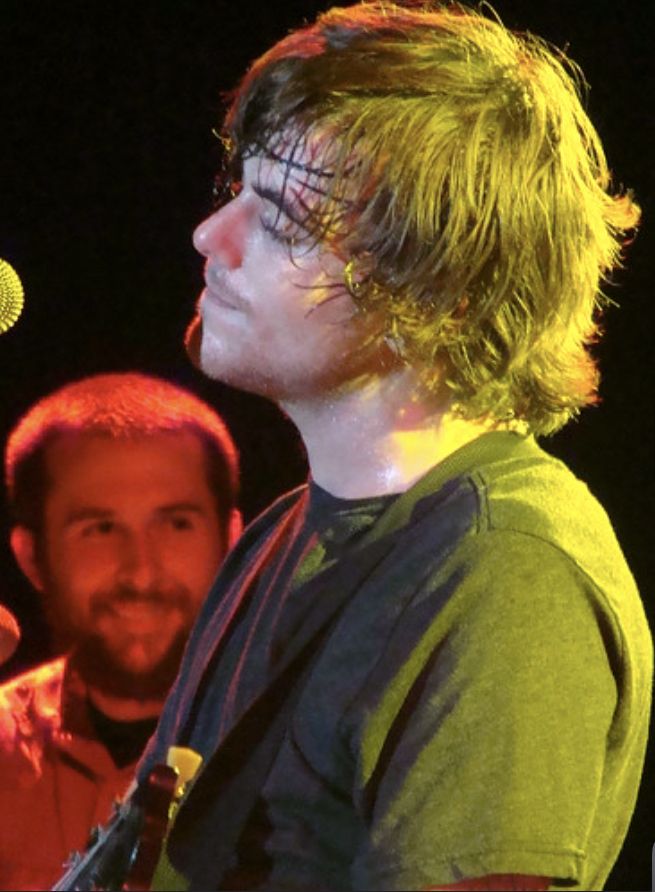 a man standing next to a microphone in front of another man holding a guitar and singing into a microphone