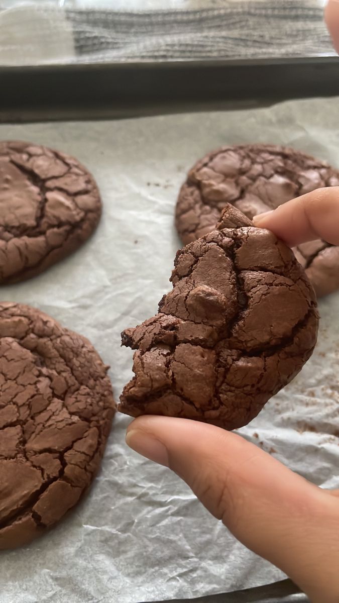 a person holding a chocolate cookie in front of other cookies on a baking sheet with wax paper