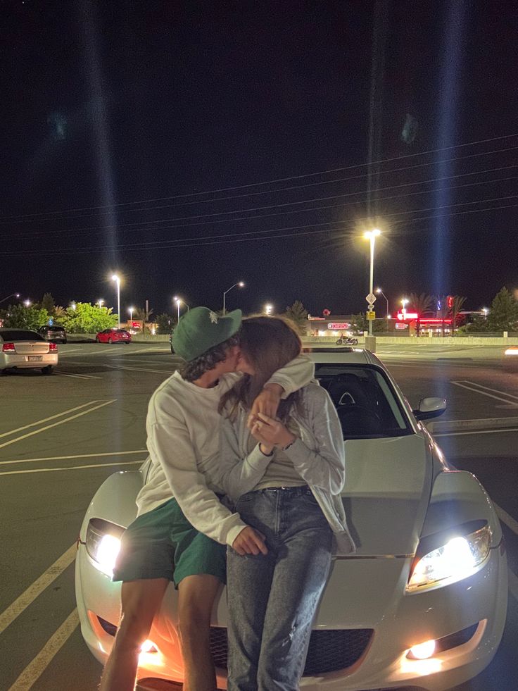 two people are sitting on the hood of a car at night, one is kissing the other's head