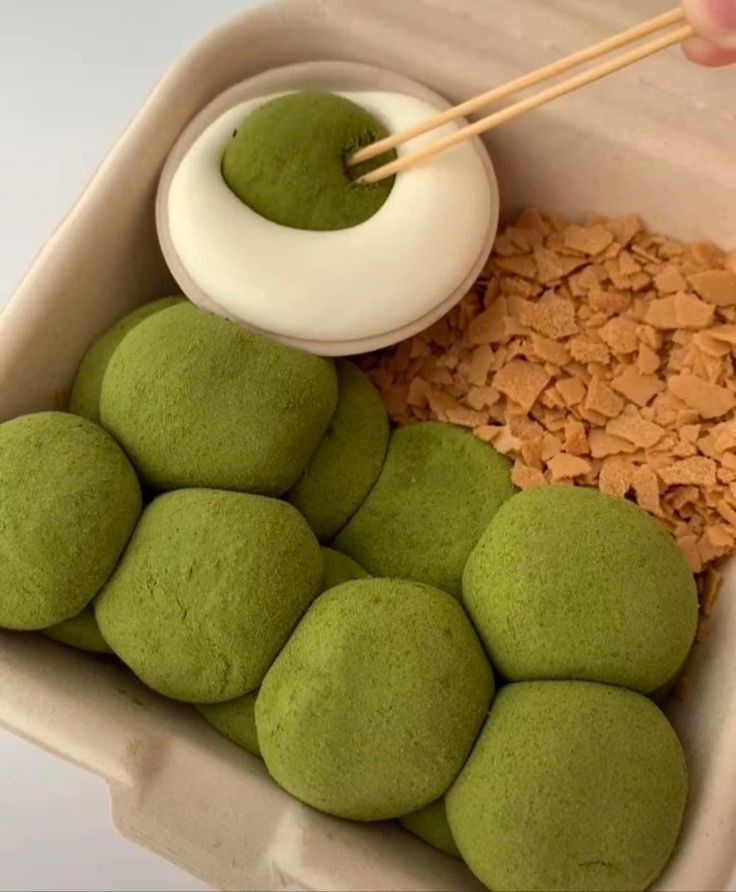 a container filled with lots of green food next to a bowl of rice and chopsticks