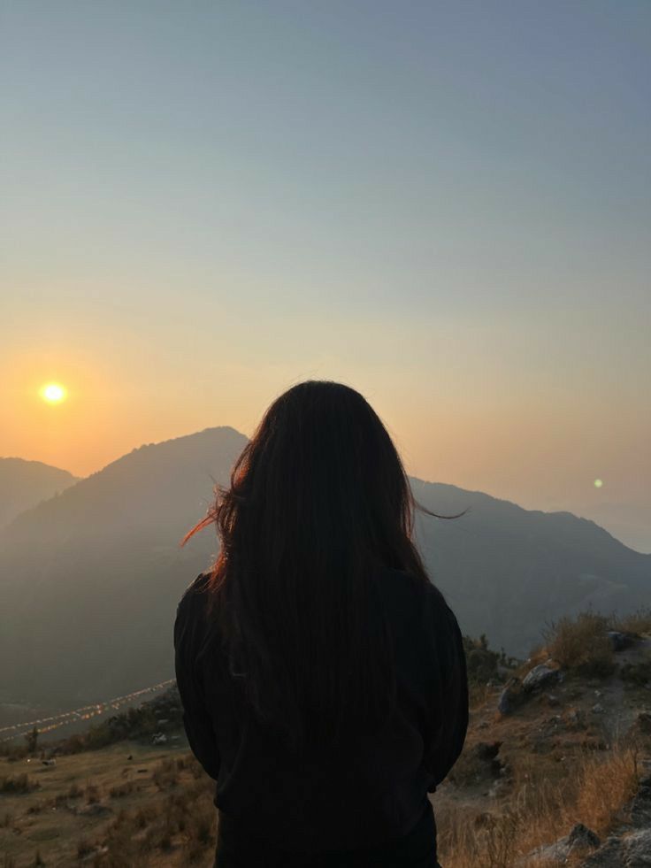 a woman standing on top of a hill looking at the sun setting in the distance