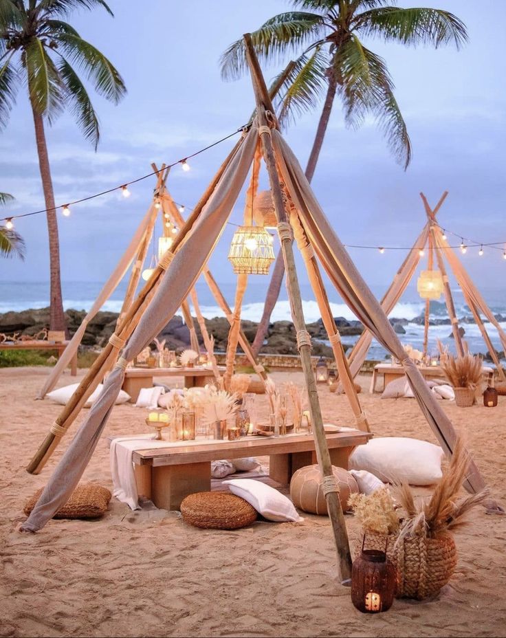 a table set up on the beach with candles and lights hanging from it's poles