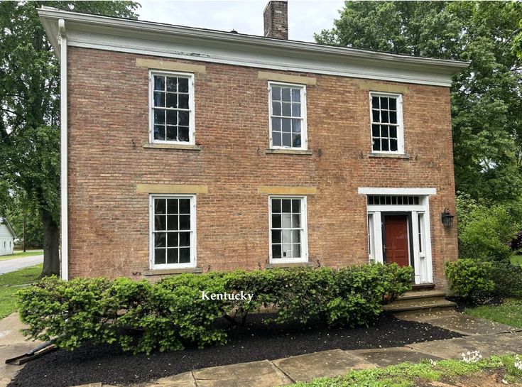 an old brick house with white trim on the front door and windows, surrounded by shrubbery