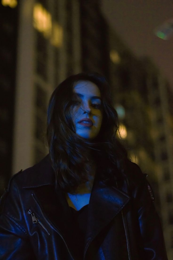 a woman with long hair standing in front of a tall building at night, wearing a black leather jacket