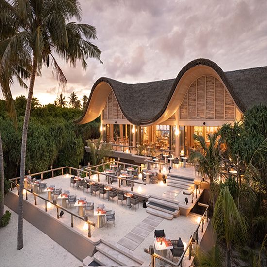 an outdoor dining area with tables and chairs on the beach at dusk, surrounded by palm trees