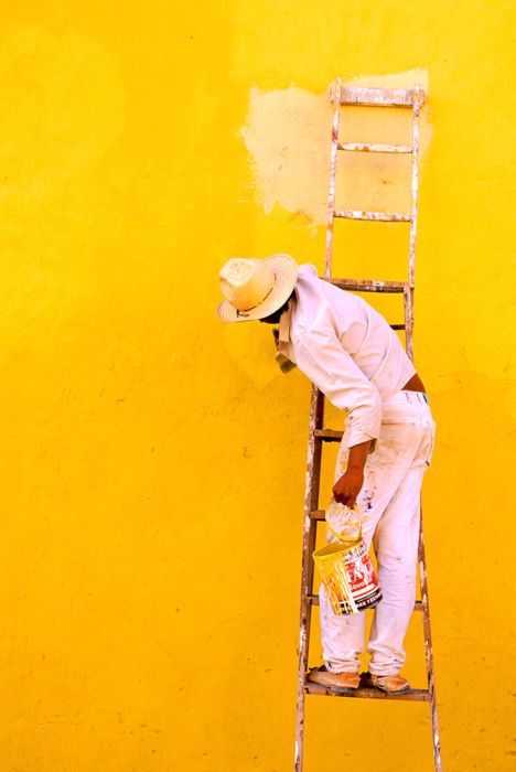 a man is painting the side of a yellow wall with a ladder and paint bucket