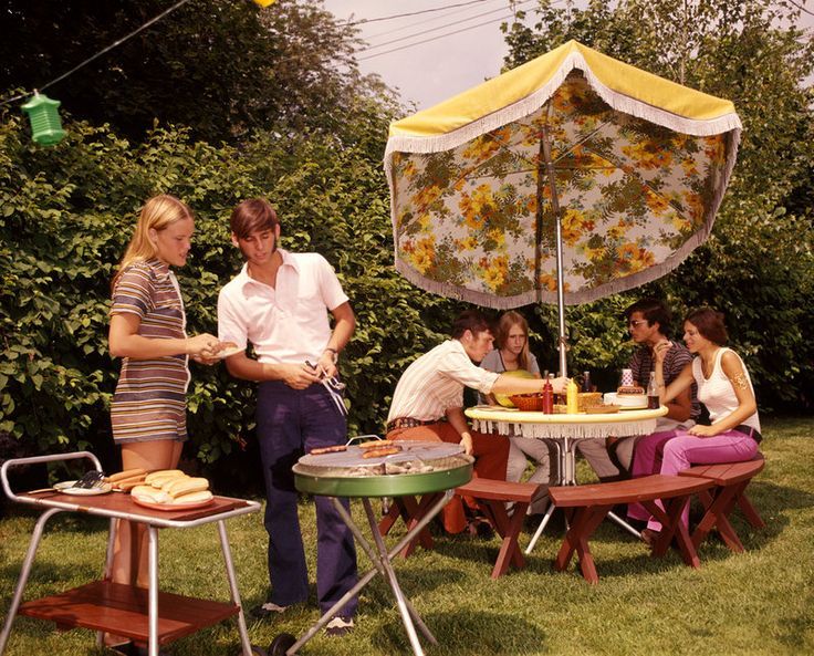 a group of people sitting around a table with an umbrella over it in the grass