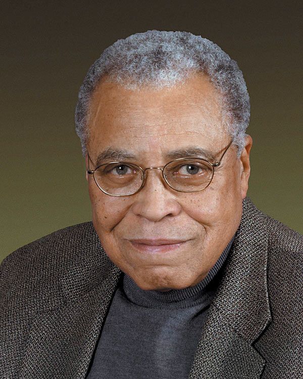 an older man wearing glasses and a gray sweater smiles at the camera with his right hand on his hip