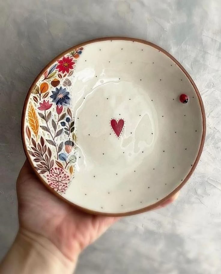 a hand holding a small bowl with flowers and hearts painted on the side, in front of a white background