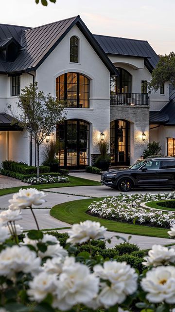 a black car parked in front of a large white house with lots of flowers around it