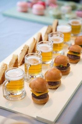 mini burgers and beer are lined up on a long tray with glasses of beer