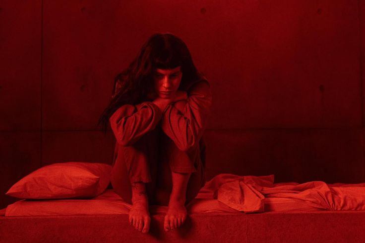 a woman sitting on top of a bed in a room with red light behind her