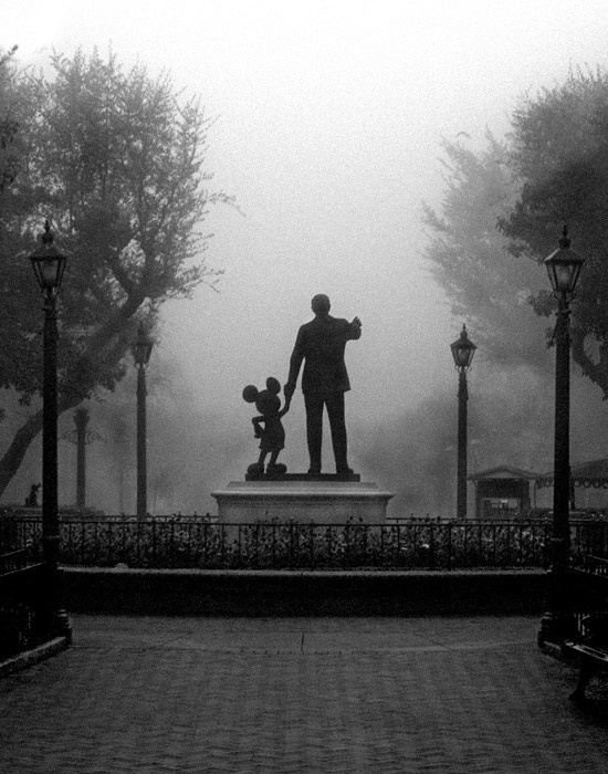 a statue of mickey and minnie mouse holding hands in front of a foggy park