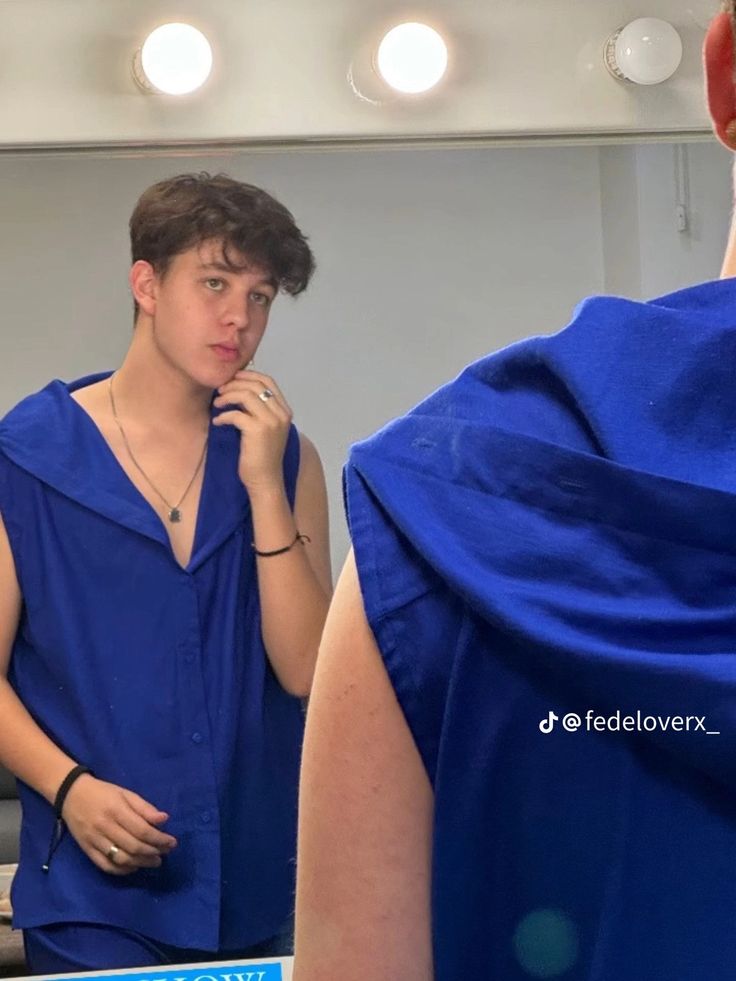 a young man is looking at himself in the mirror while brushing his teeth and wearing a blue shirt