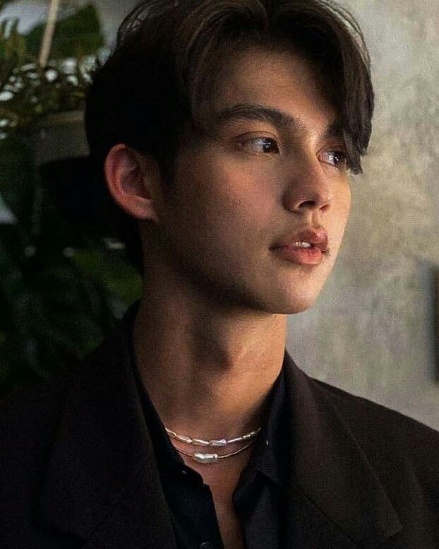 a young man wearing a black shirt and silver necklace looking off into the distance while standing in front of a potted plant