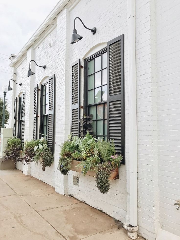 a white brick building with black shutters and window boxes filled with plants on the outside