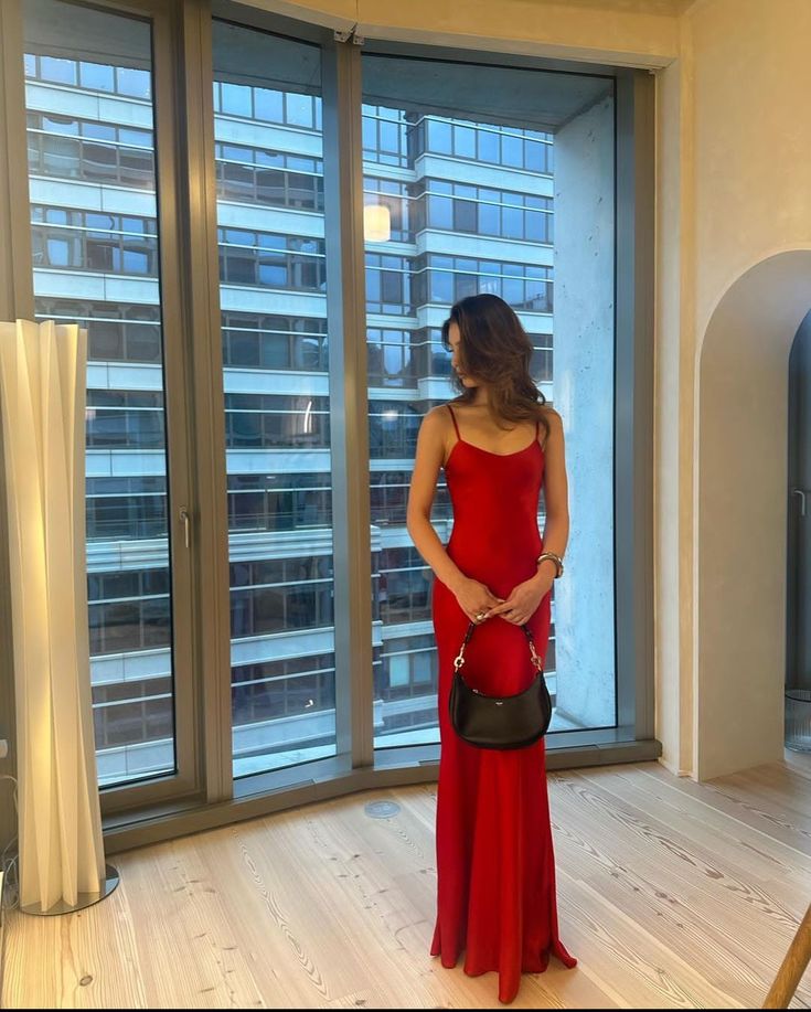 a woman in a red dress standing next to a window and holding a black purse