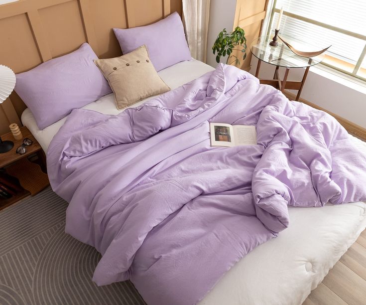 an unmade bed with purple comforter and pillows in a small room next to a window