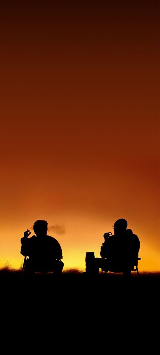 two people sitting in chairs on top of a field at sunset with the sun setting behind them