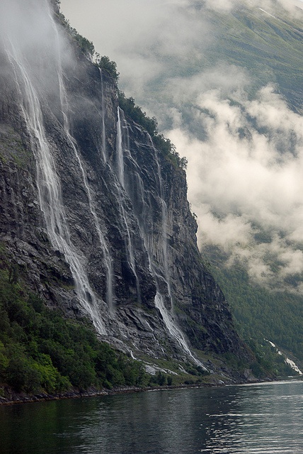 a boat is in the water near a mountain with waterfalls on it's side