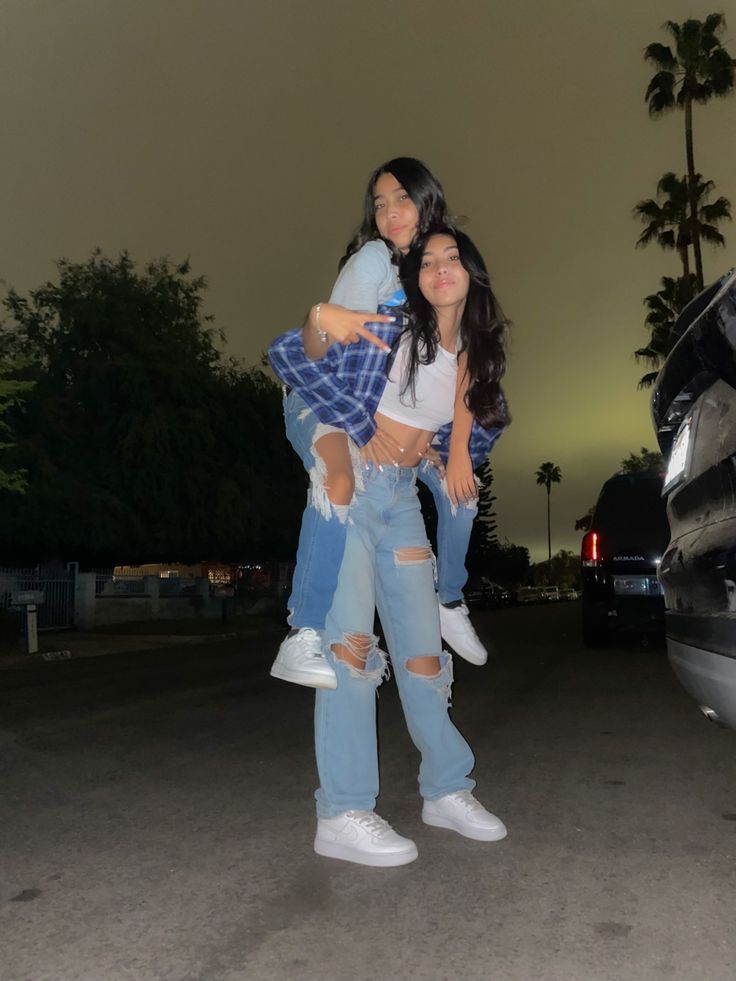 two women standing in the middle of a parking lot at night with palm trees behind them