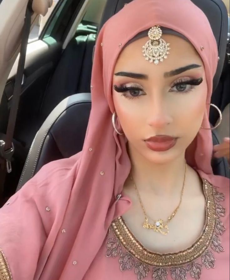 a woman wearing a pink hijab and gold jewelry in front of a car