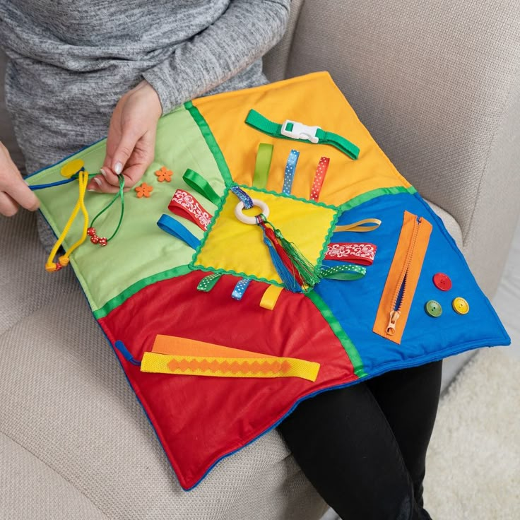 a woman sitting on a couch holding a colorful play mat with various items in it