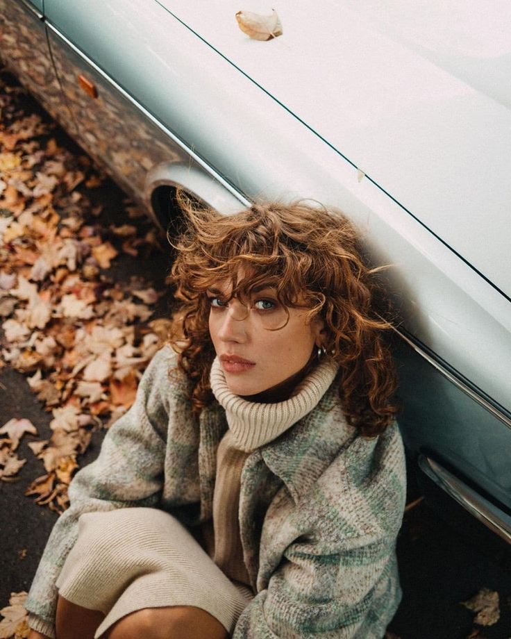 a woman sitting on the ground next to a car with leaves all over it and her hair blowing in the wind