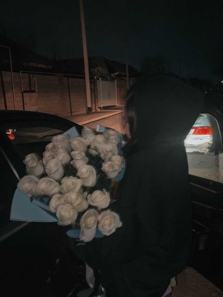 a person in a black hoodie holding a bunch of white roses next to a car