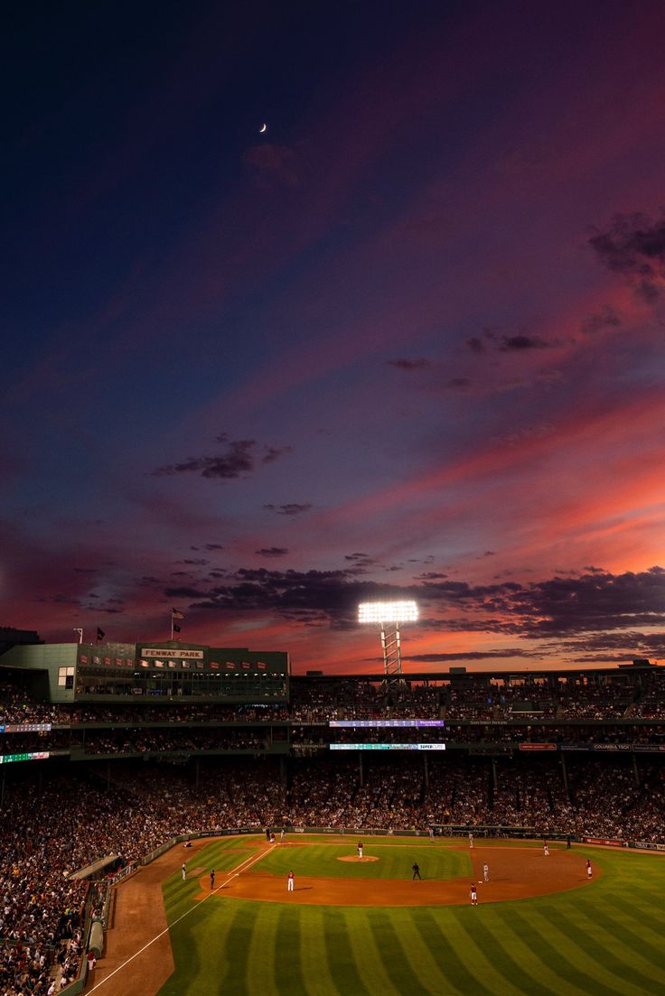 a baseball field at night with the sun setting in the distance and clouds lit up