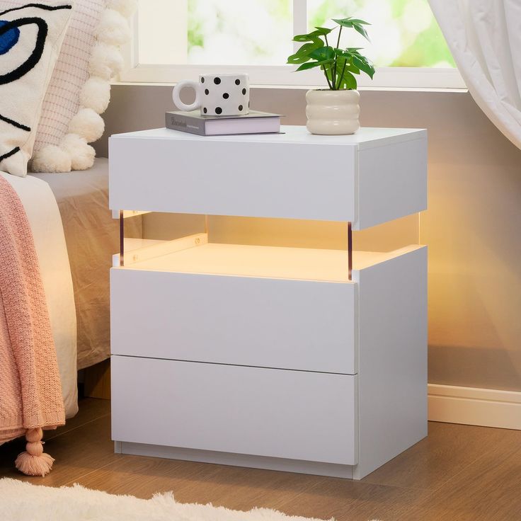 a white nightstand sitting next to a bed with a plant on top of the night stand