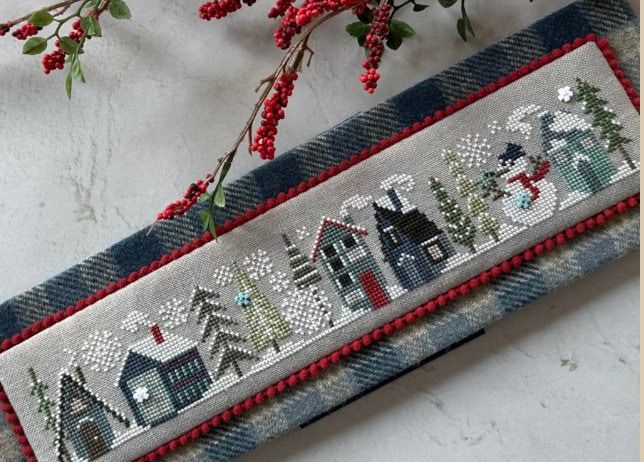 a cross stitch christmas banner hanging on a wall next to a branch with red berries