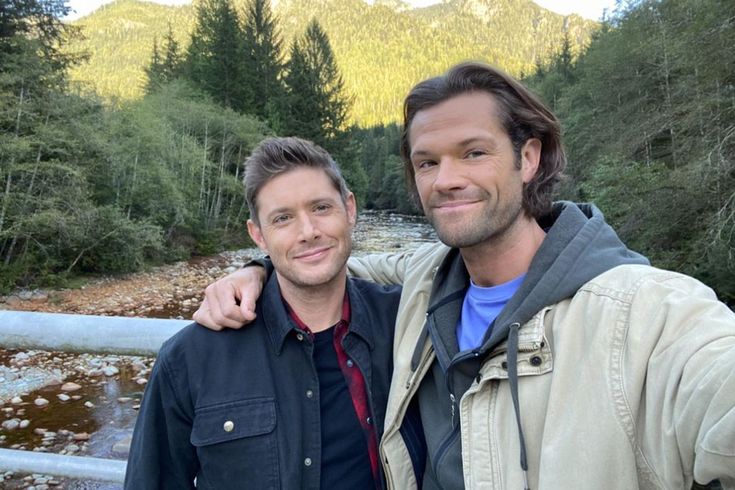 two men standing next to each other in front of a river and mountains with trees