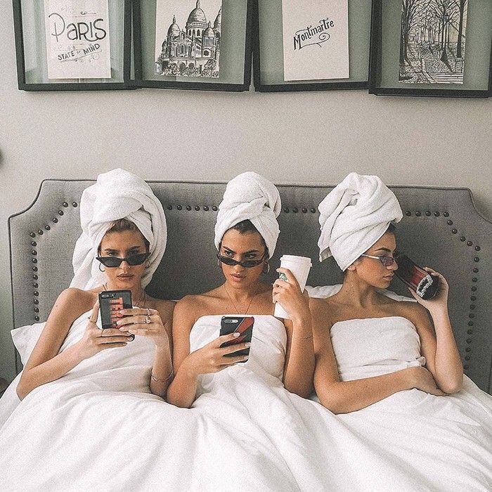 three women laying in bed with towels wrapped around their heads looking at cell phones and drinking coffee