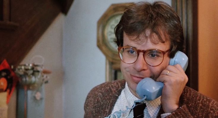 a man with glasses talking on a phone in his living room while wearing a sweater and tie