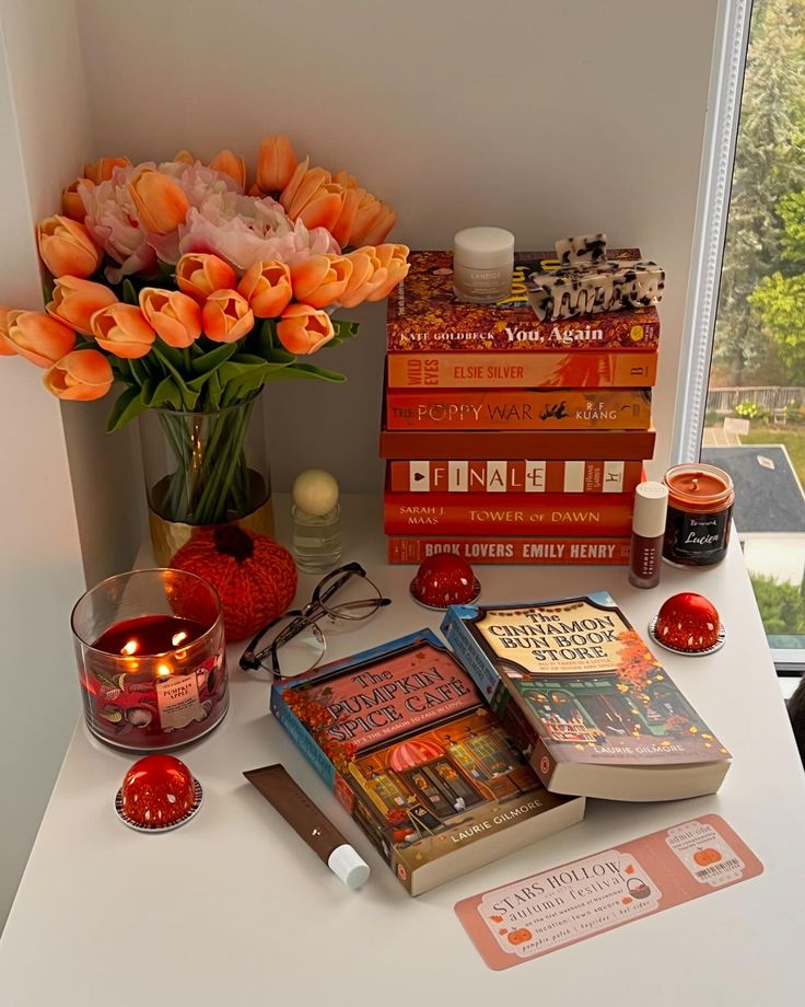 an assortment of books and candles on a white table next to a vase with orange flowers