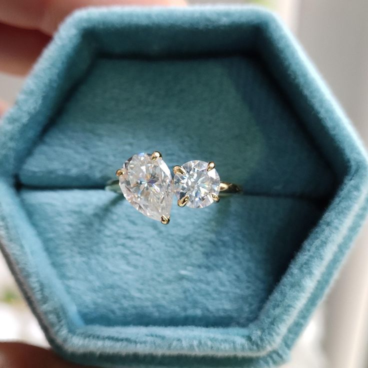 an engagement ring with two pear shaped diamonds sits in a blue velvet box on top of a window sill