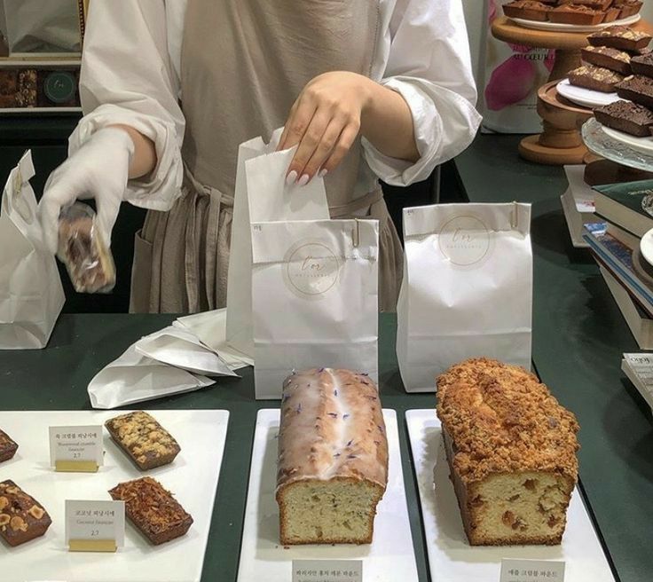 a woman in white shirt standing next to counter filled with cakes and pastries on trays