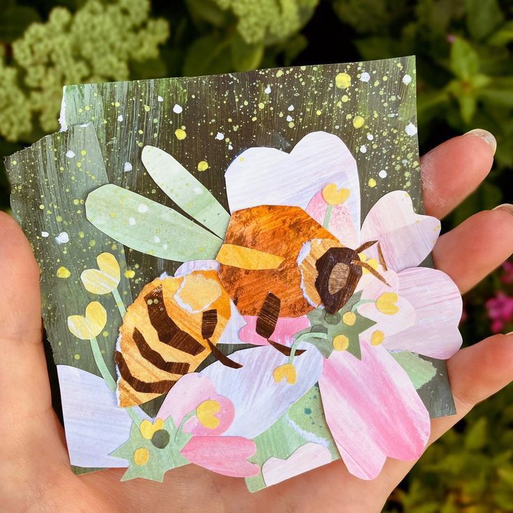 a hand holding up a piece of paper with a painting of a bee on it