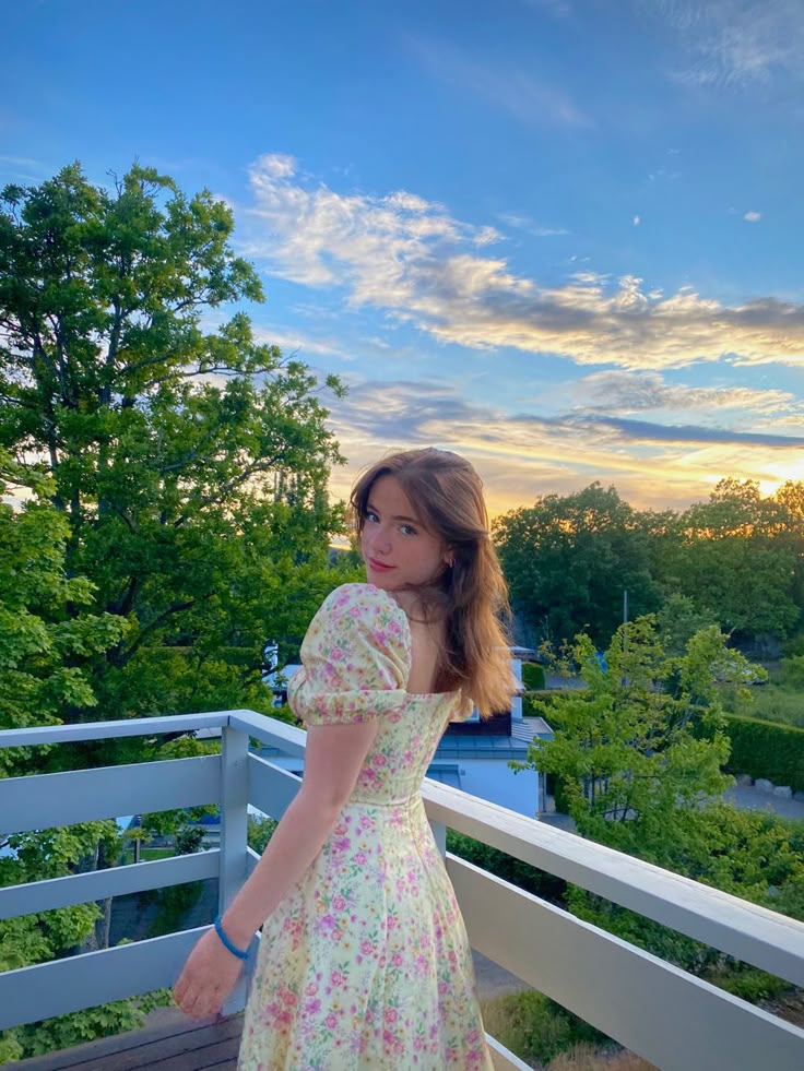 a woman in a dress is standing on a balcony looking at the camera with trees and sky in the background