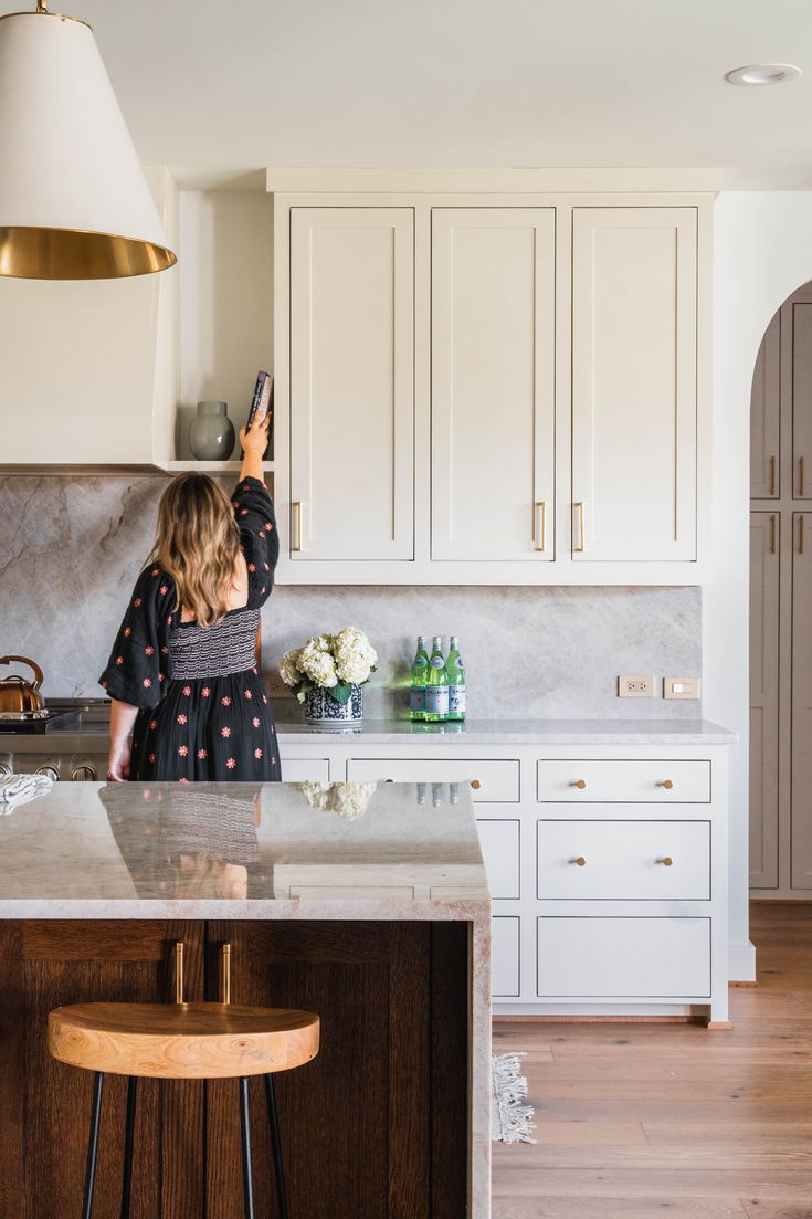 a woman standing in a kitchen next to a counter with flowers on it and holding a cell phone