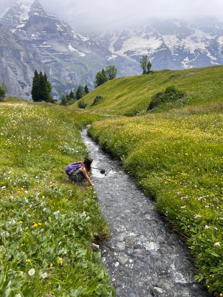 a woman kneeling down next to a stream in a field with mountains in the background