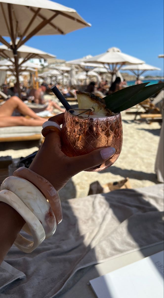 a person holding a drink in their hand on the beach with umbrellas and lounge chairs behind them