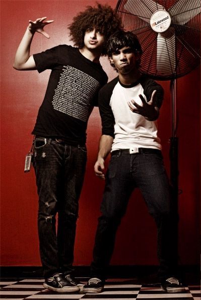 two young men standing next to each other in front of a wall with a fan