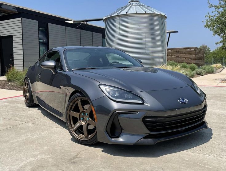 a gray sports car is parked in front of a building with silos and water tanks