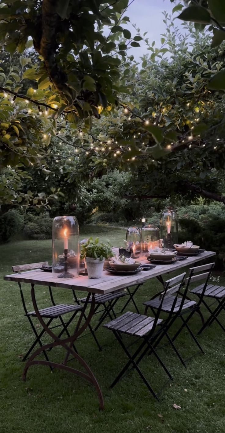 an outdoor dining table set up with candles, plates and glasses on it in the grass